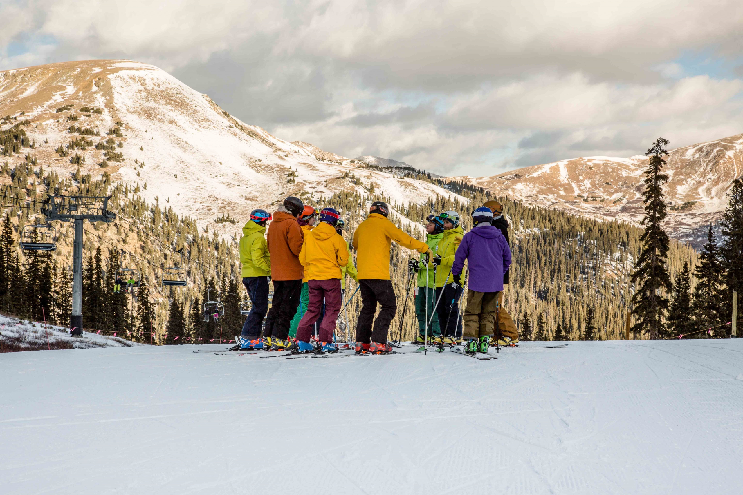 A group of alpine and telemark skiers gathers in a circle at the top of a run during the afternoon alpenglow