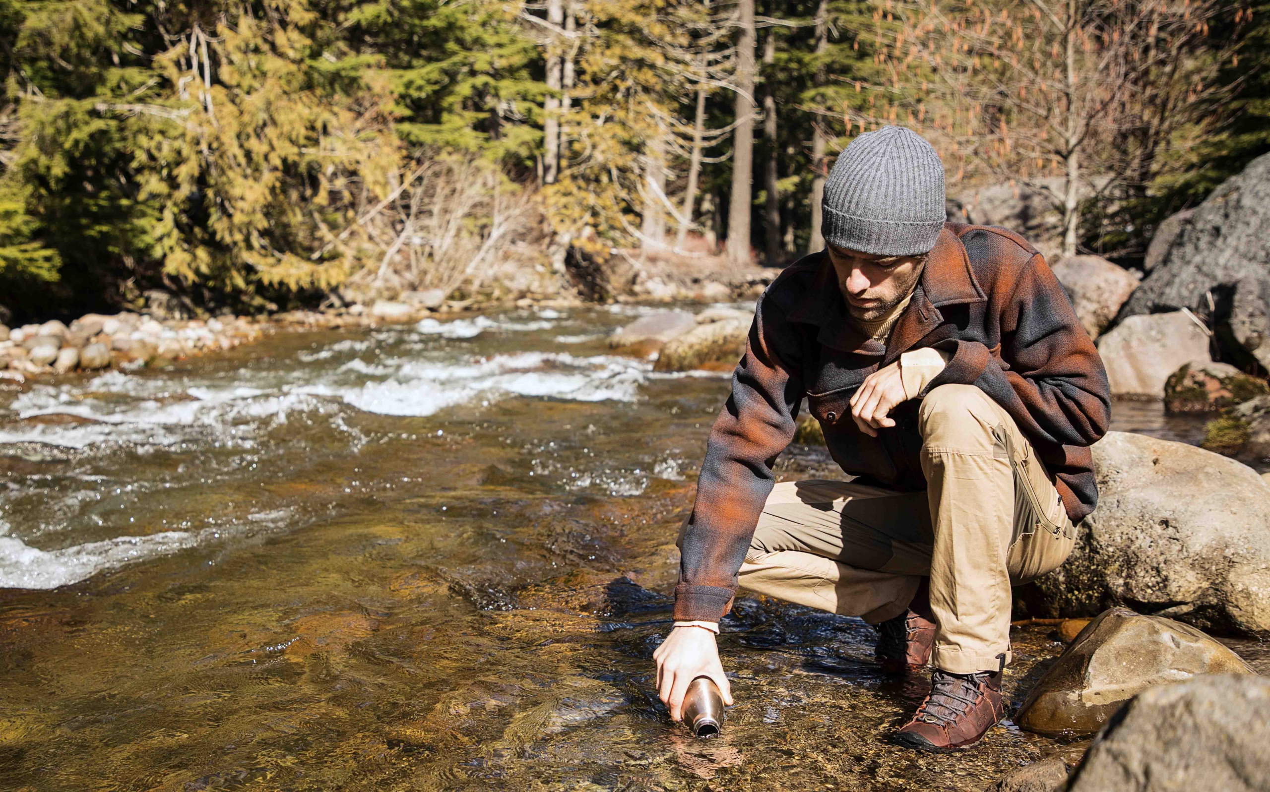 A man in a flannel and beanie stands by a stream in his KEEN boots and fills a water bottle