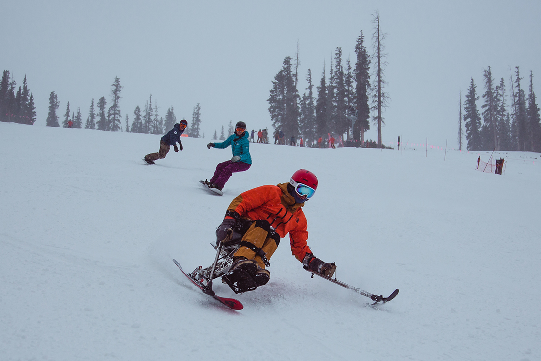 An adaptive skier descends a ski run ahead of two snowboarders